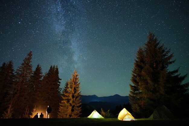 Silhouette of hikers resting besides burning bonfire near illuminated tourist tents on camping site in dark mountains under night sky with sparkling stars. Active lifestyle and outdoor living concept.