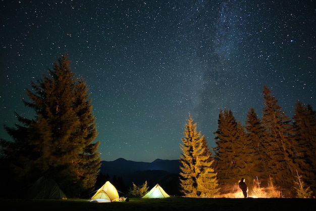 Silhouette of lonely hiker resting besides burning bonfire near illuminated tourist tents on camping site in dark mountains under night sky with stars. Active lifestyle and outdoor living concept.