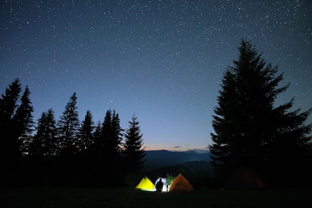 Silhouette of lonely hiker resting besides burning bonfire near illuminated tourist tents on camping site in dark mountains under night sky with stars Active lifestyle and outdoor living concept