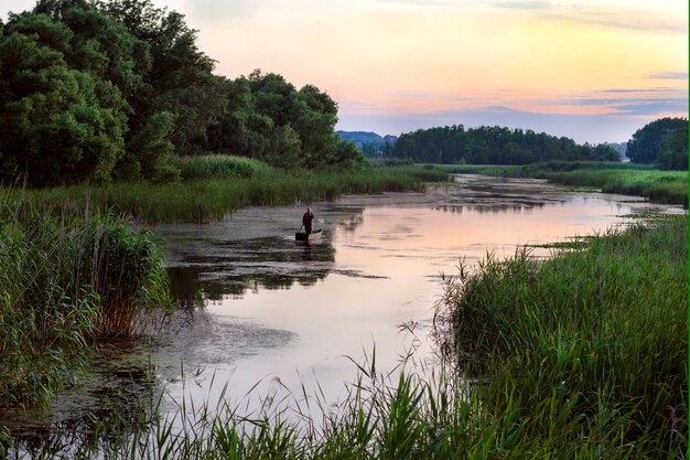 Silhouette of a man in a boat with an oar moving along a winding river overgrown with reeds, in the evening.