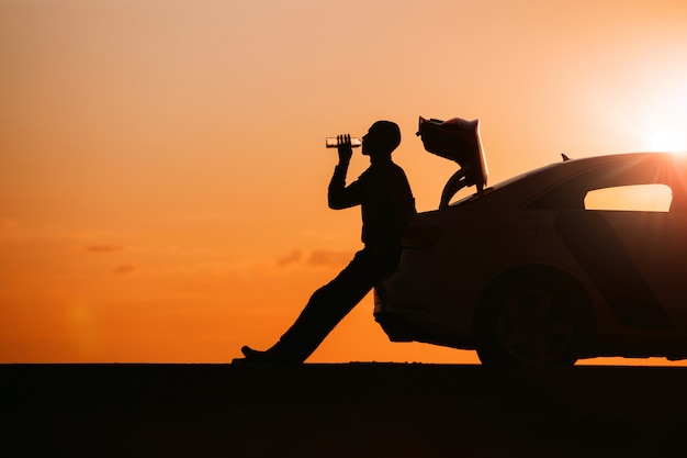 Silhouette of man driver relaxing after a ride, sitting on the trunk of his car and drinking water from a bottle