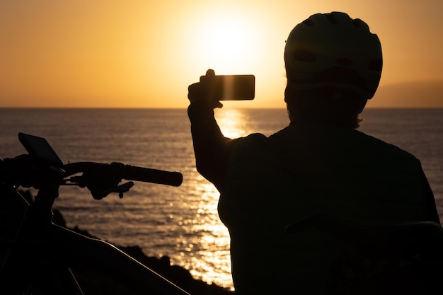 Silhouette of man looking at sunset taking a photography with mobile phone, wearing sport helmet after cycling along the sea.