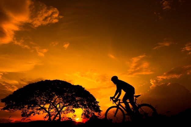 Silhouette Man riding Uphill with Bicycle at sunset with orange sky in countryside. 