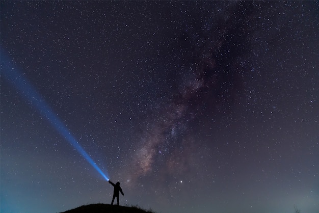 Silhouette of a man with a flashlight, observing beautiful, wide blue night sky with stars and Milky way galaxy. Astronomy, orientation, clear sky