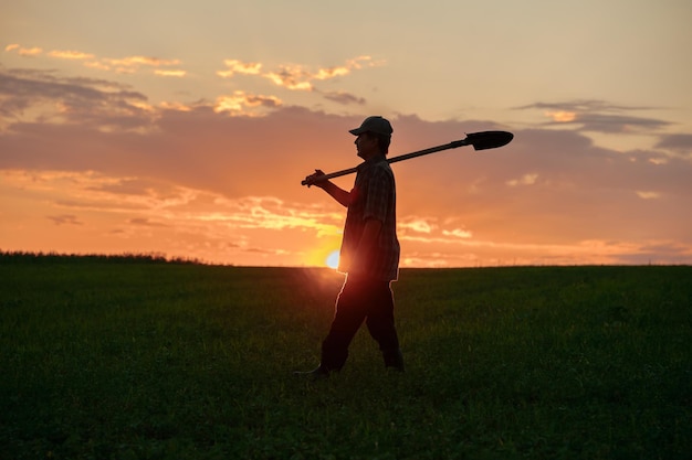 Silhouette of the man with the shovel walking through the field at sunset worker in agricultural fie