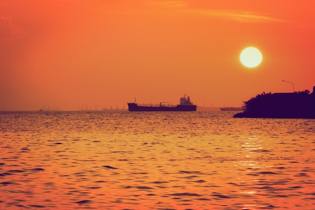 Silhouette of marine cargo ship is anchored by the sea at sunset