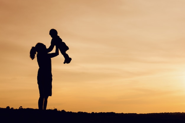 Silhouette of mother and daughter lifting child in air over scenic sunset sky at riverside. 