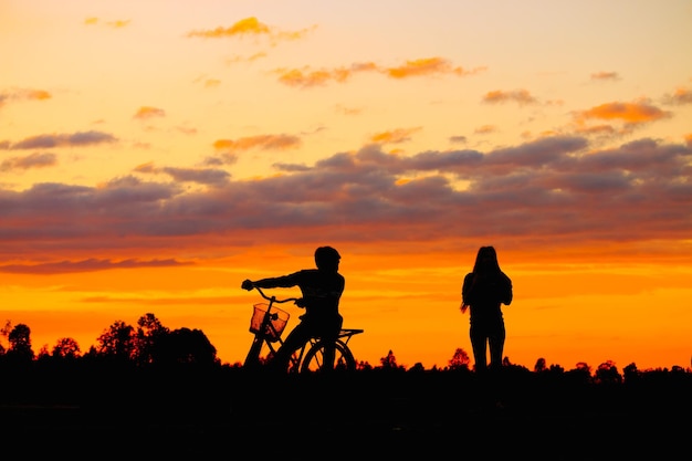 Silhouette person with bicycle by woman standing on field against sky during sunset