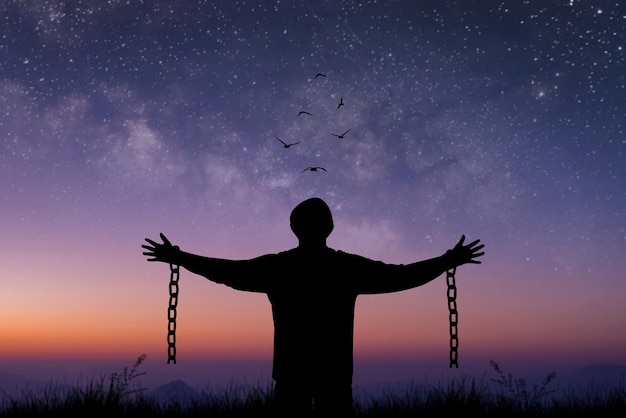 Silhouette prisoner stood with his arms outstretched with a chain broken in his hand standing alone on top of mountain at night time with star Milky Way and bird over the sky