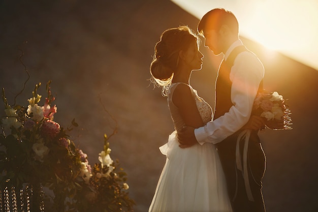 Silhouette sweethearts at sunset. Beautiful and fashionable brunette model girl with modeling hairstyle in white lace dress posing at the sunset with handsome men in stylish vest and white shirt