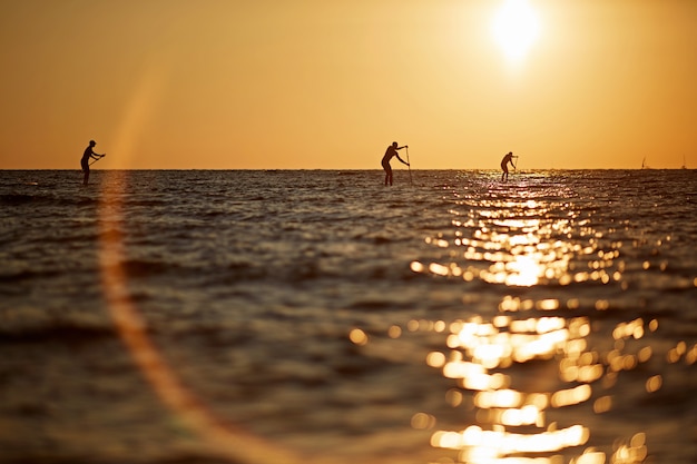 Silhouette of three young people rowing on a surfboard to the horizon in the open sea beautiful scenic sunset