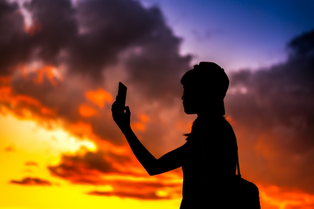 Silhouette of a tourist woman holding phone taking pictures with dramtic sunset sky background