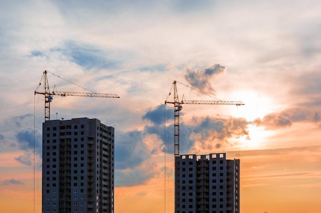 Silhouette of tower cranes and unfinished multistorey high buildings under construction site in the rays of the setting sun of beautiful sunset