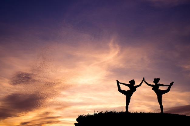 Silhouette of two Beautiful girls Yoga Standing Bow Pulling Pose on the mountain with  beautiful sunset sky.