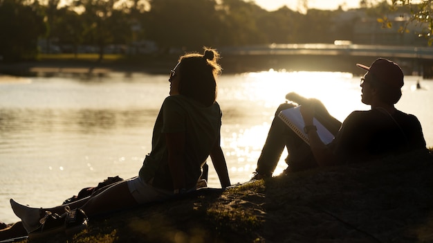 Silhouette of Two Friends on the riverside Playing Guitar on Park in a Sunny Day. Vacations Concept