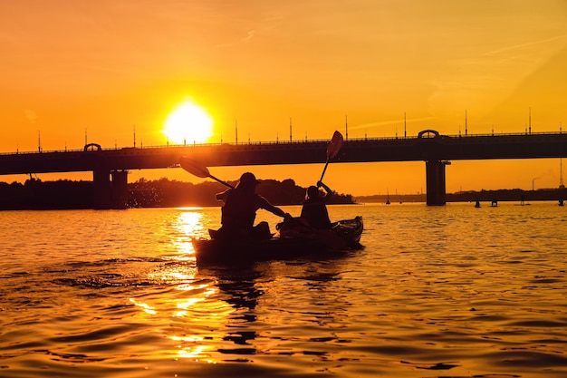 Silhouette of two persons kayaking on city river at sunset Small local travel with family