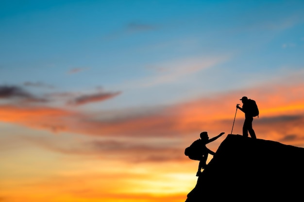 Silhouette two with backpack is climbing a cliff at sunrise He had an effort to climb all the way to the top They help each other to reach the top of the mountain