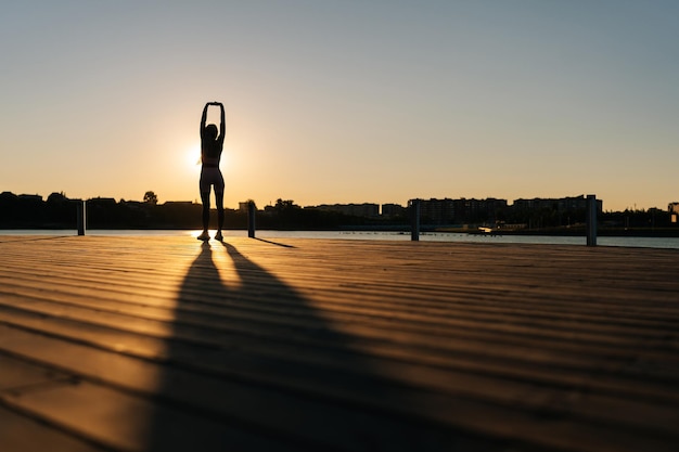 Silhouette of unrecognizable athletic woman in sporty outfit warm up muscles practicing stretching exercises before running by water in city park