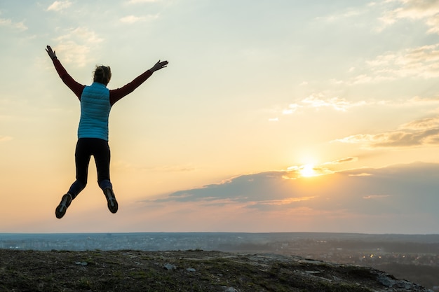 Silhouette of a woman hiker jumping alone on empty field at sunset in mountains. Female tourist raising her hands up in evening nature. Tourism, traveling and healthy lifestyle concept.