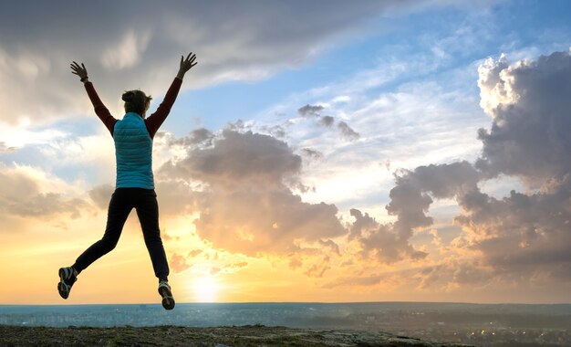 Silhouette of a woman hiker jumping alone on empty field at sunset in mountains.