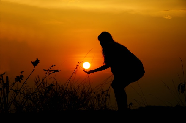 The silhouette of a woman is holding the sun Has a sunset background