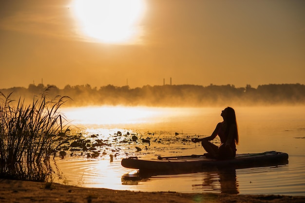 Silhouette of a woman in the lotus position on an inflatable SUP board at dawn and the horizon with mists on the water atmosphere of relaxation