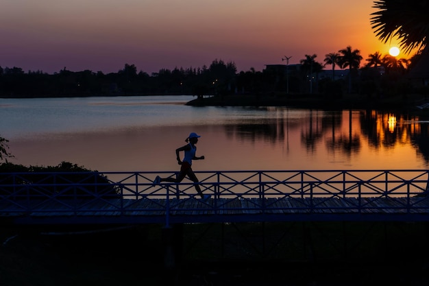 Silhouette woman running across the bridge at lakeside against sunset