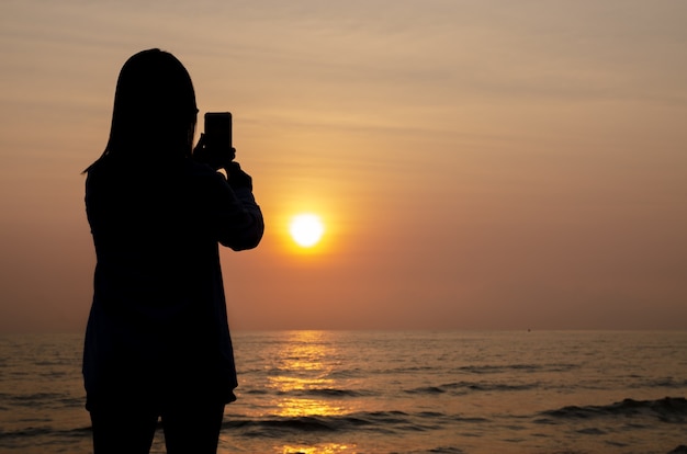 Silhouette of woman take photo sunset on the sea and colorful sky by smartphone.