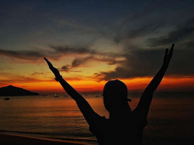 Silhouette woman with arms raised at beach against sky during sunset