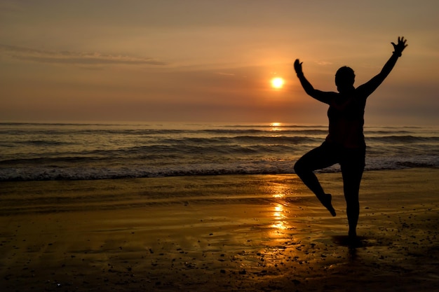 Silhouette of a woman with open arms and on one foot on the shore of a beach at sunset