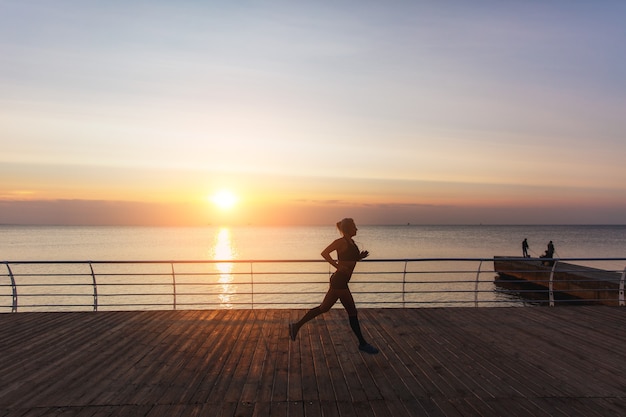 The silhouette of a young beautiful athletic girl with long blond hair in headphones, who listens to music and runs at dawn over the sea