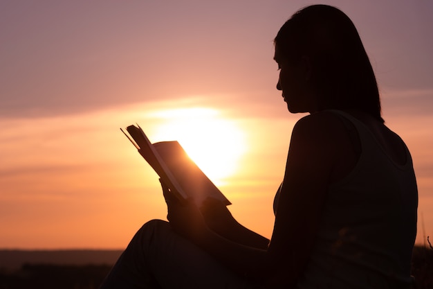 Silhouette of a young beautiful woman at dawn sitting on the ground and carefully staring at the open book