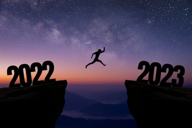 Silhouette of young businessman jumping over a cliff from 2022 to 2023 with beautiful night sky star and Milky Way over on the sky Concept of planning goal challenge new year resolution