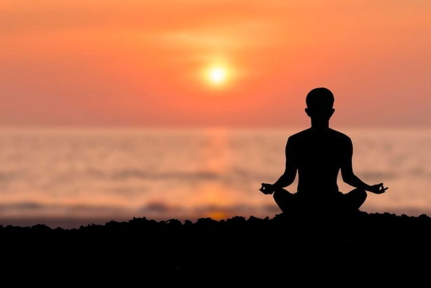 Silhouette of young female sitting practices yoga and meditating in lotus position alone on the rock in the morning at the sea with beautiful sunrise and orange sky She felt calm and happy