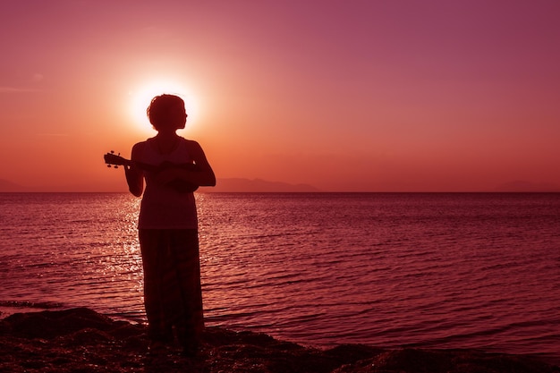 Silhouette of young girl standing on the beach with ukulele at the sunset against the sun