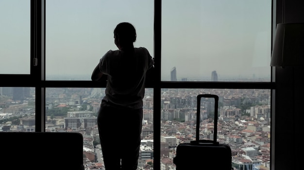 Silhouette of young girl traveler with suitcase in hotel room with panoramic city view. She is standing near the window and dancing.