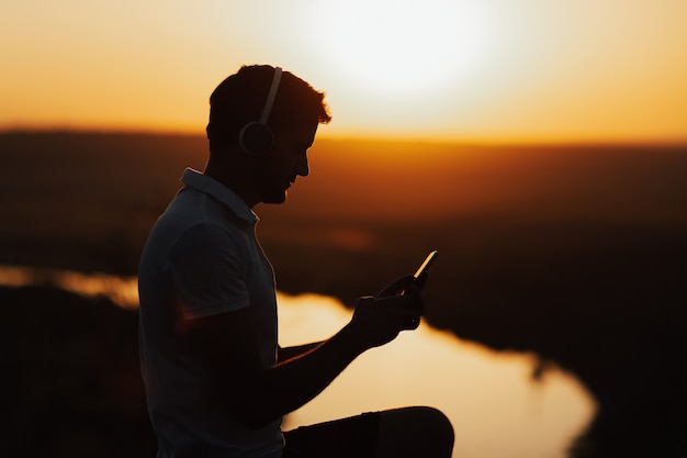 Silhouette of young man holding mobile phone. He enjoying sunset and listening to the music on the smartphone.