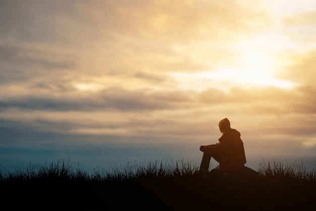 Silhouette of young man sits praying alone at the top of the mountain at sunset with beautiful natural sunlight