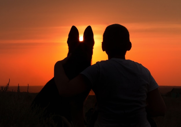 Silhouette of a young man with a dog enjoying beautiful sunset in a field, boy fondle his favorite pet on nature, concept friendship of animal and human