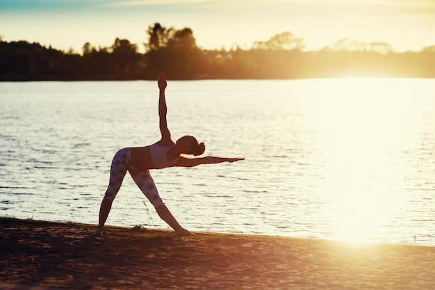 Silhouette of a young woman doing yoga exercises on the lake beach at sunset