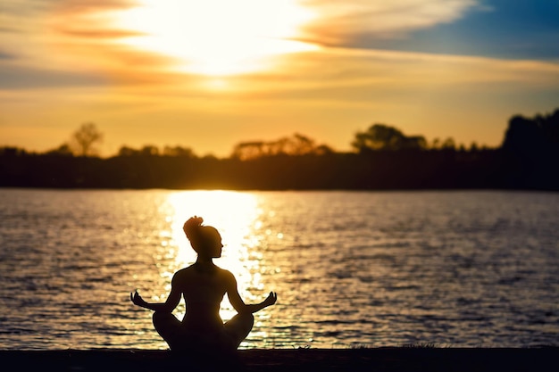 Silhouette of a young woman doing yoga exercises on the lake beach at sunset