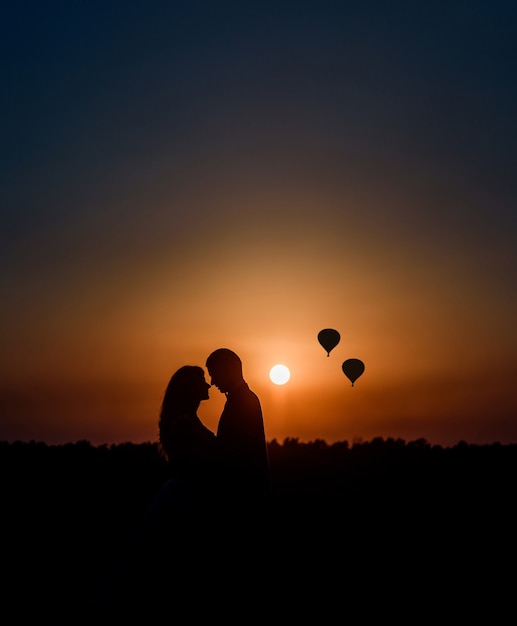 Silhouettes of a couple hugging each other before a sunset and air balloons up in the sky