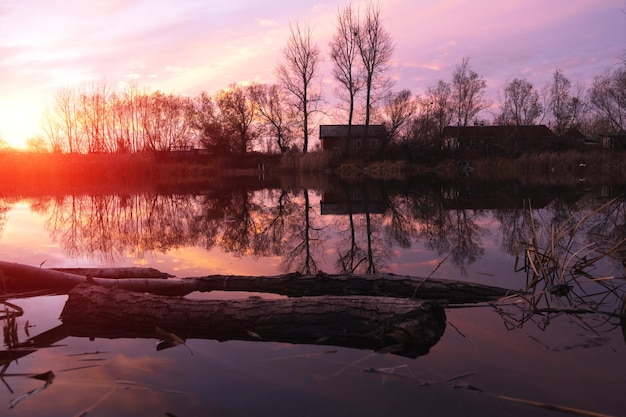 Silhouettes of houses and trees of an abandoned village on the lake against a bright scarlet, autumn sunset.