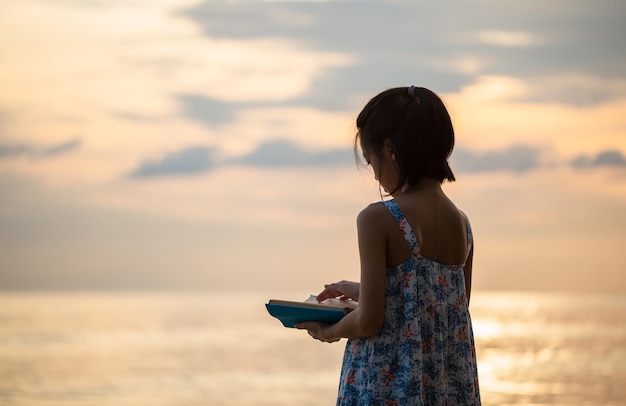 Silhouettes of little cute girl playing on the beach, Kid girl in dress reading a book on the beach sunset evening sky