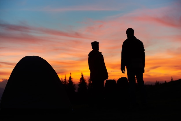 Silhouettes of a man and woman standing near their tent on top of a mountain stunning sunset 