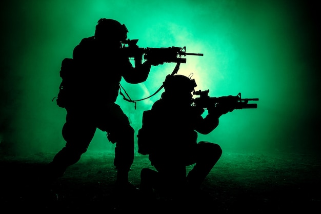 Silhouettes of two army soldiers, U.S. marines team in action, surrounded fire and smoke, shooting with assault rifle and machine gun, attacking enemy with suppressive gunfire during offensive mission