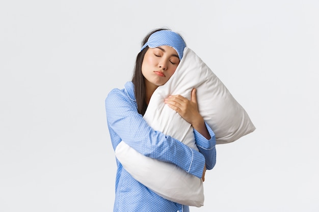 Silly and cute asian girl in blue sleeping mask and pajamas, hugging pillow tight as unwilling wake-up, dont want get up from bed in morning, heavy sleeper posing over white background.