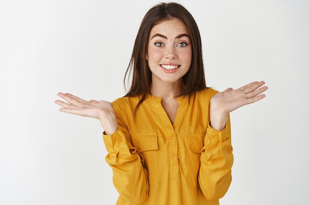 Silly and cute young woman shrugging with hands spread sideways and awkward smile, saying sorry as being clueless, standing over white wall