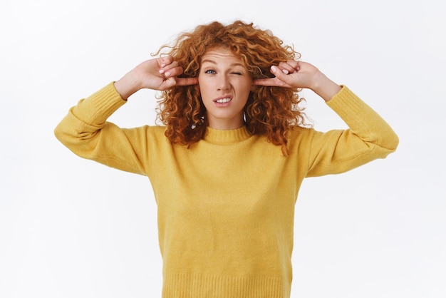 Silly displeased cute redhead curly girl shut her ears plug fingers into earholes and squinting cringe from hearing annoying noise disturbed by loud sound siren standing white background