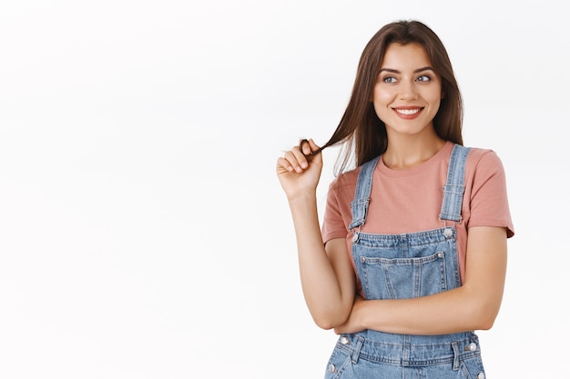 Silly, dreamy alluring brunette woman in dungarees, t-shirt rolling hair strand on finger and smiling happy, looking away as recalling, imaging something good, standing white background pleased